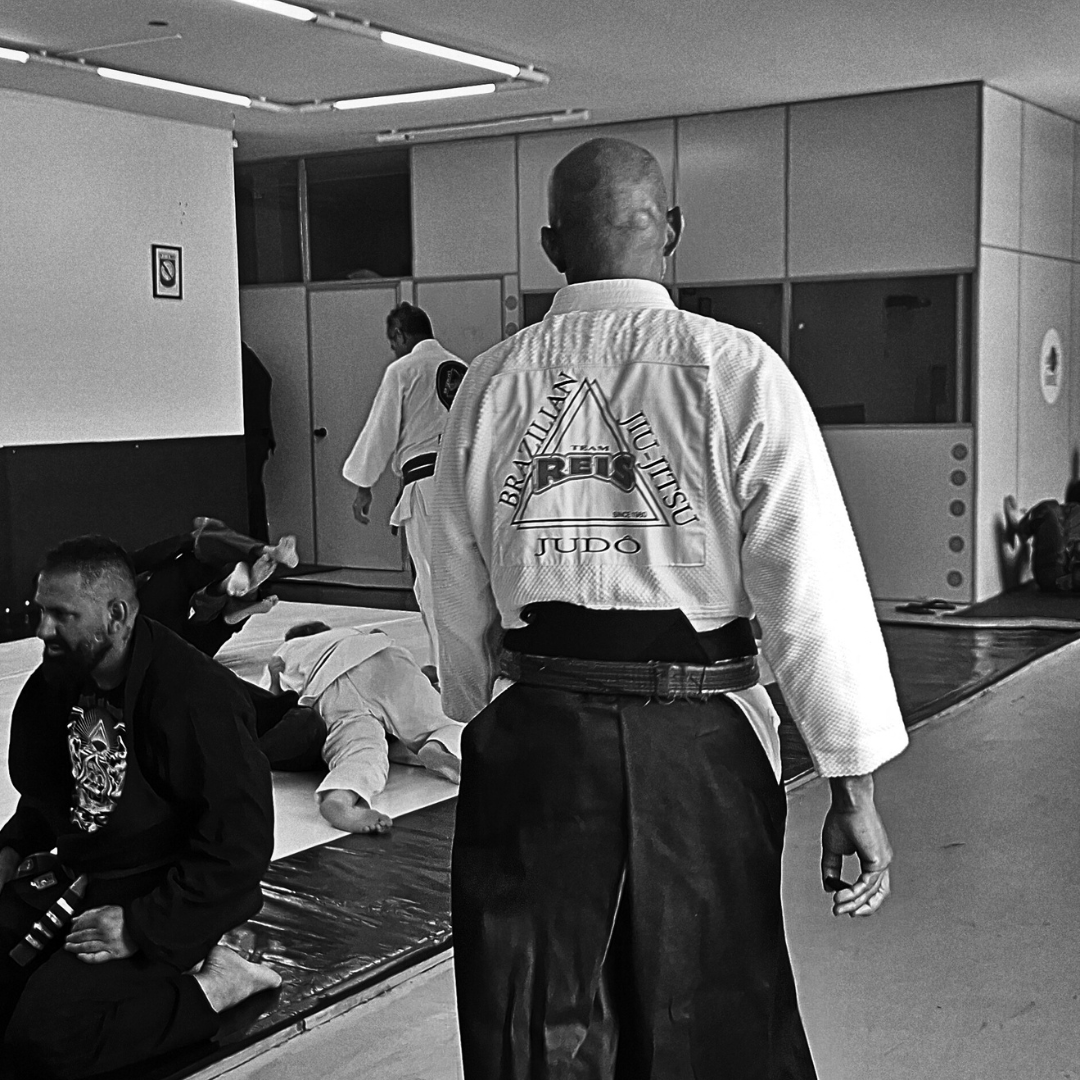 Mestre Reis de costas no tatame, com o logo da academia no kimono